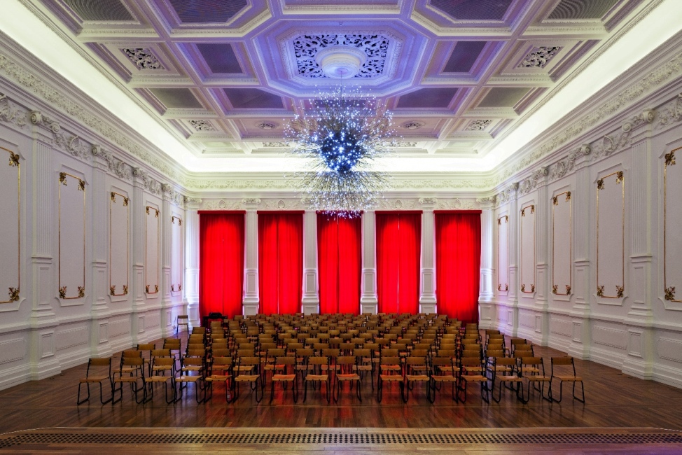 Uma sala branca, com paredes ornamentais, muitas cadeiras dispostas ao meio e um lustre grande azul na parte central da sala