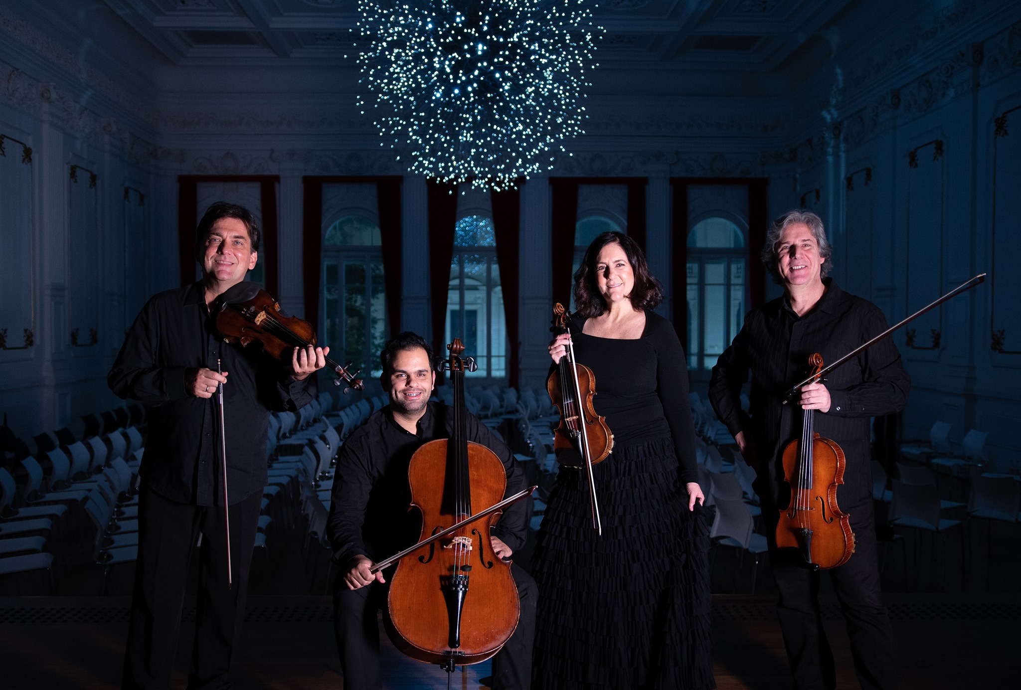 Da direita para esquerda um homem de roupa preta segurando uma viola, uma mulher de preto segurando um violino, um homem sentado de preto segurando um violoncelo e um homem de pé segurando um violino