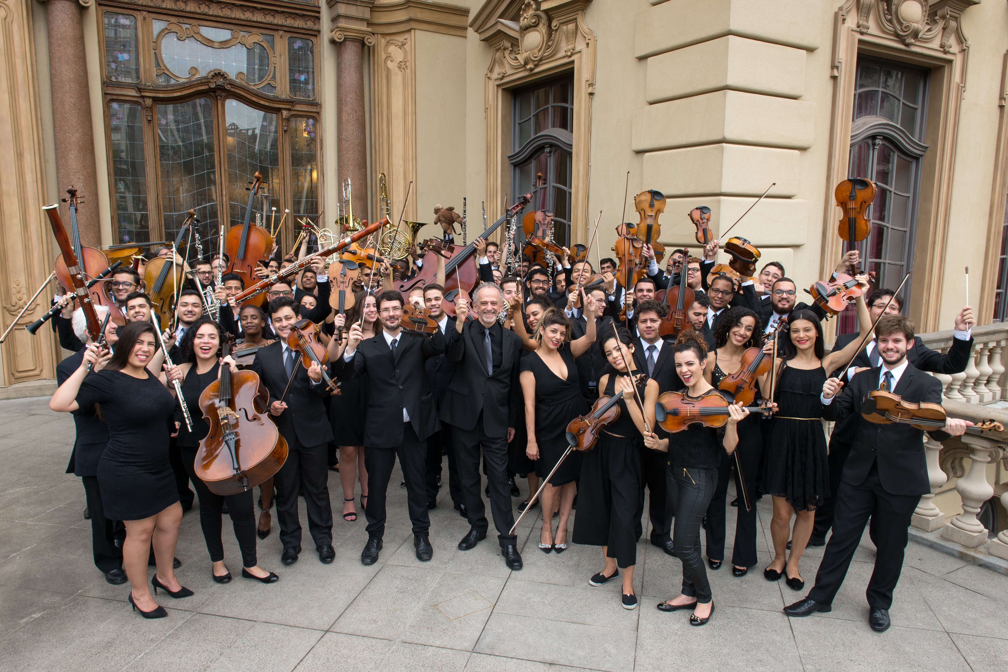 Uma foto com uma orquestra completa na fachada de um prédio, todos os músicos estão de preto com seus instrumentos para cima.