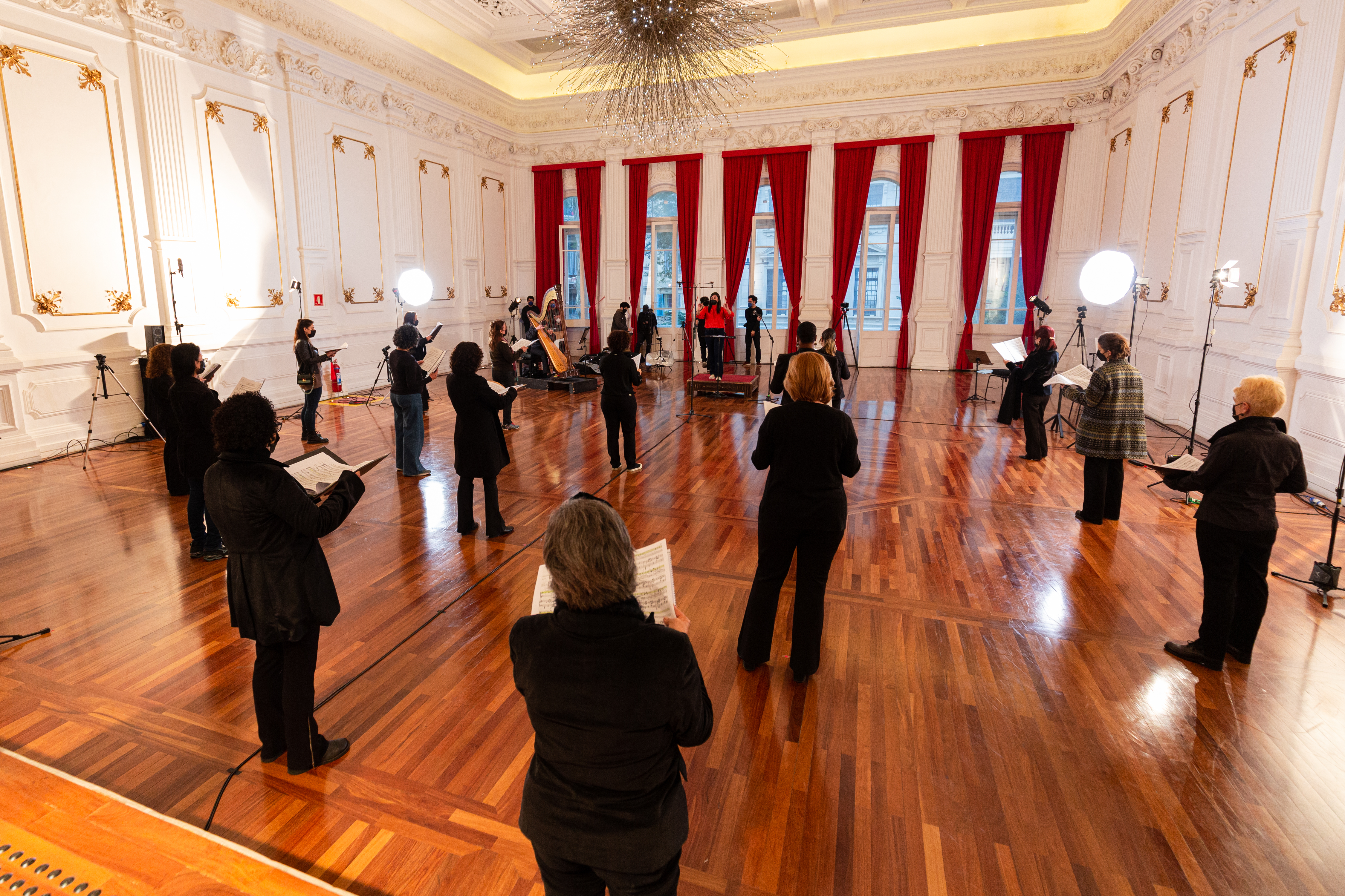 Integrantes do Coral Paulistano com roupas pretas e segurando partituras, dispostos na sala do conservatório. Luzes estão dispostas na sala e o ambiente esta bem iluminado e sem cadeiras.