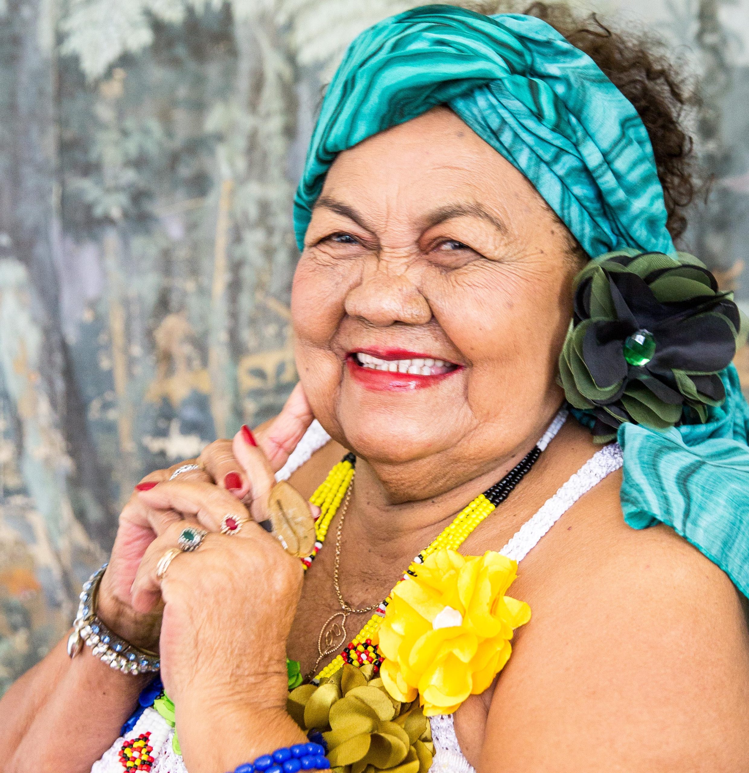 Dona Onte usa lenço azul na cabeça e flores amarelas na roupa. Ela sorri e junta as mãos.