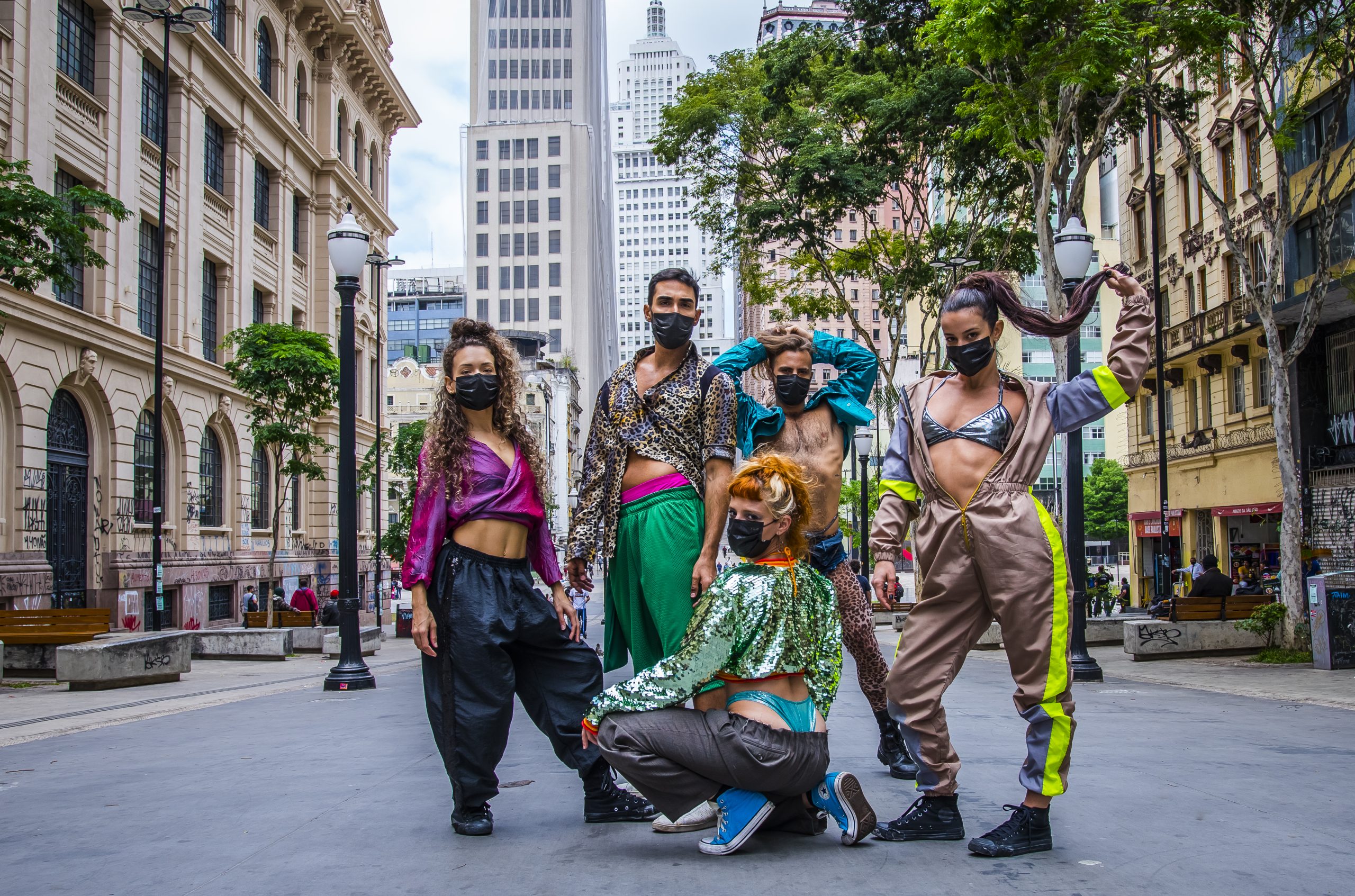 Foto que mostra cinco bailarinos do Balé da Cidade com figurinos alegres e chamativos em tons de verde, azul, rosa, amarelo e vermelho, todos olham para a câmera e estão numa praça aberta com prédio antigos nas laterais