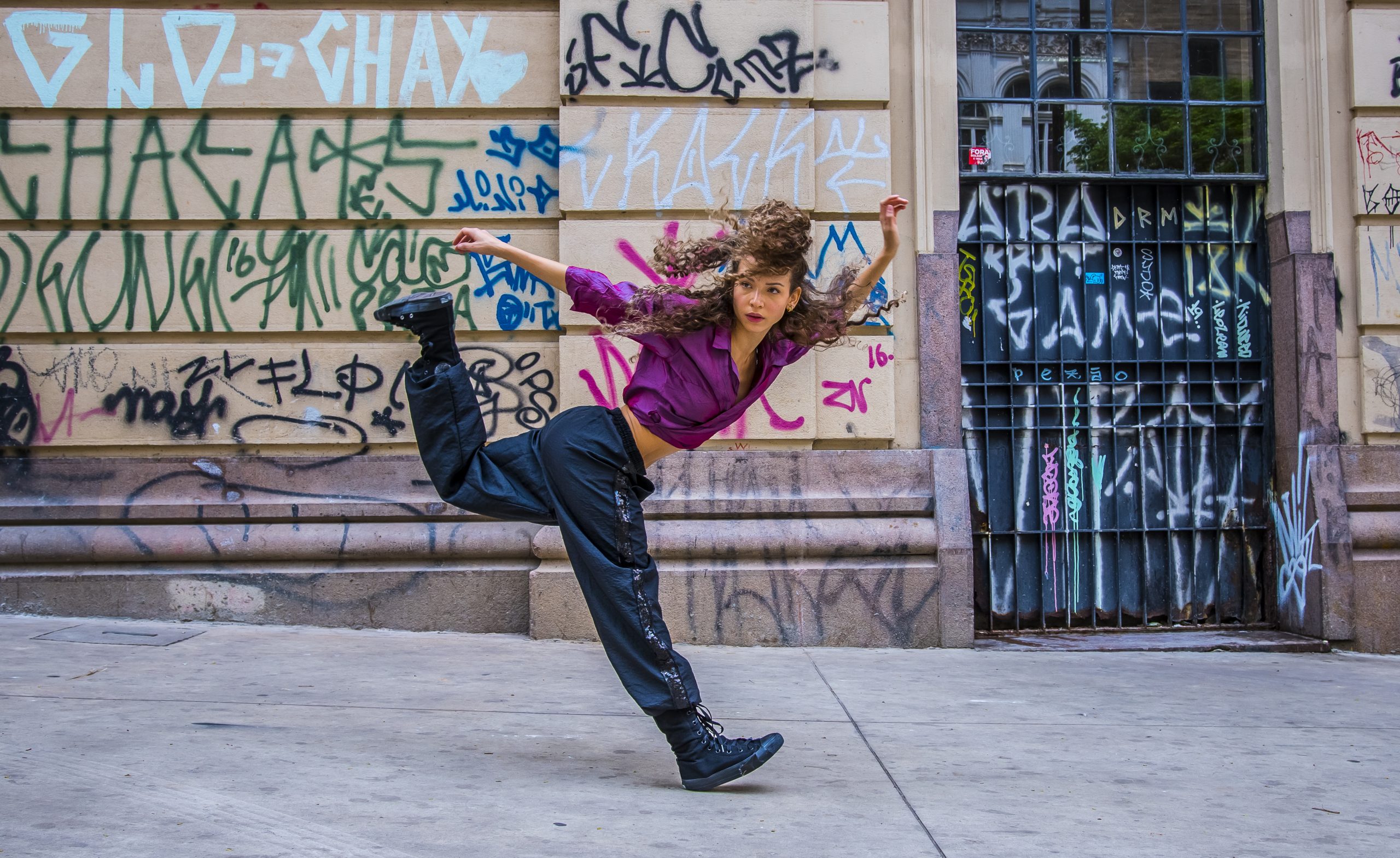 Na rua, com muros pixados, bailarina que integra o Balé da Cidade dança com calça escura e blusa de manga comprida roxa. A bailarina tem cabelos cacheados castanhos e compridos e pele branca.