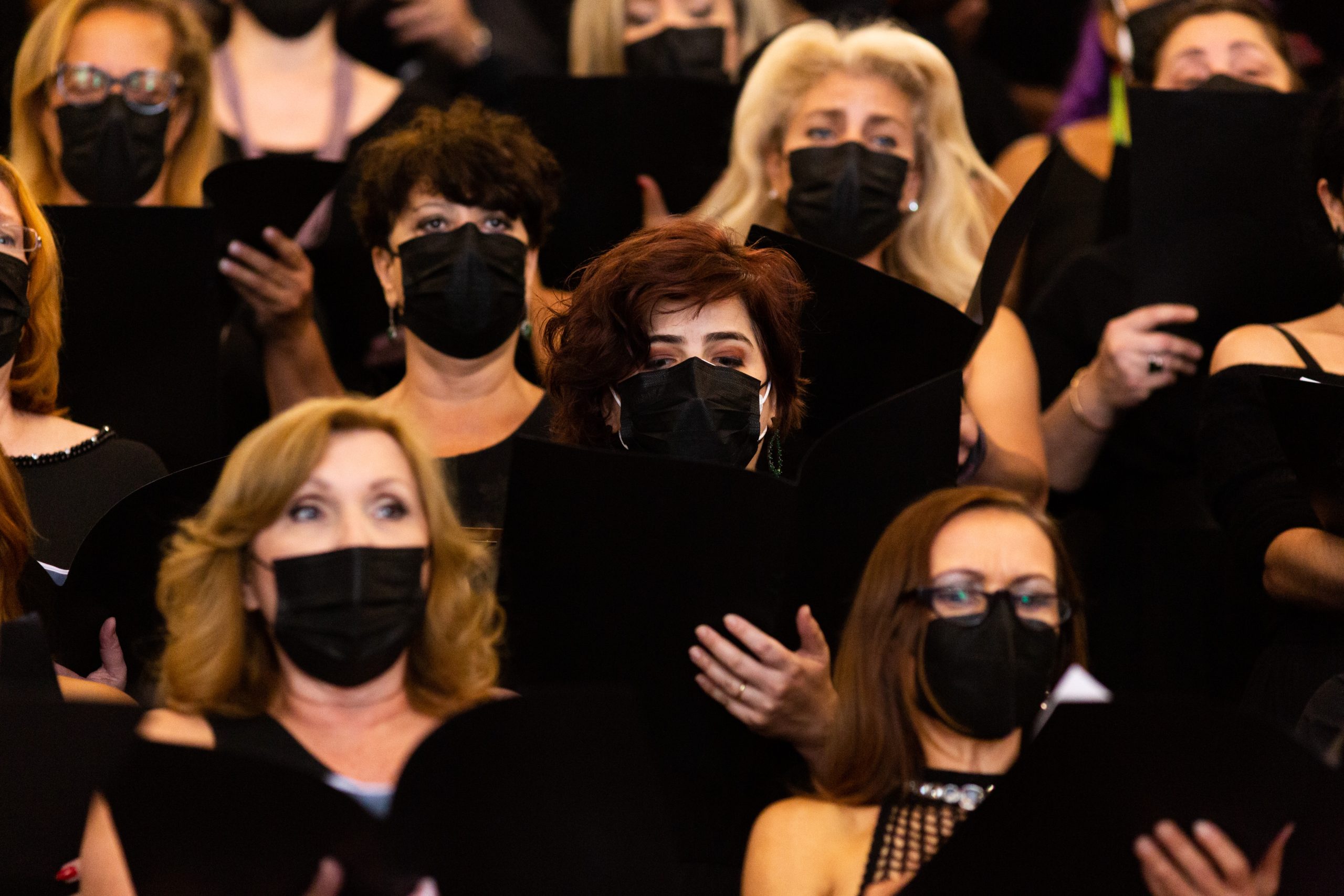 Mulheres integrantes do coro cantam segurando uma pasta na cor preta