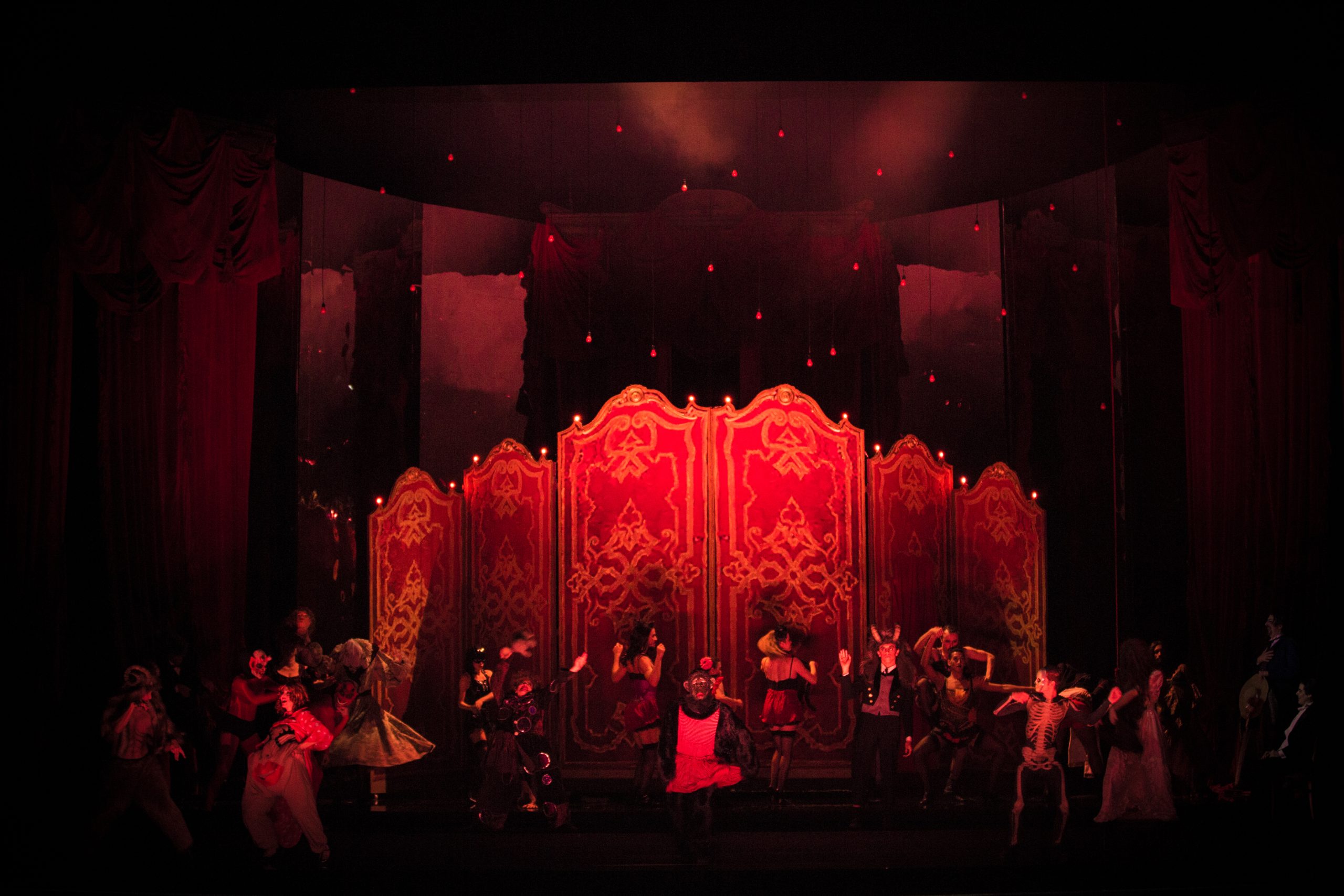 Diversos atores estão em cena, alguns com figurinos de caveiras e outros com lingeries, num cenário vermelho que remete um cabaré