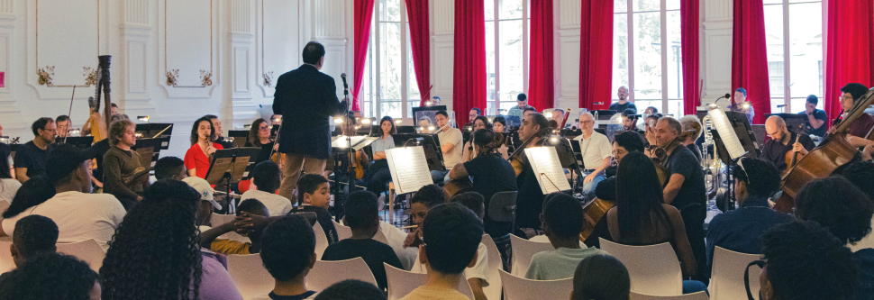 Foto de uma sala no theatro municipal repleta de músicos com um regente em pé no centro