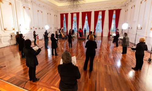 Integrantes do Coral Paulistano com roupas pretas e segurando partituras, dispostos na sala do conservatório. Luzes estão dispostas na sala e o ambiente esta bem iluminado e sem cadeiras.